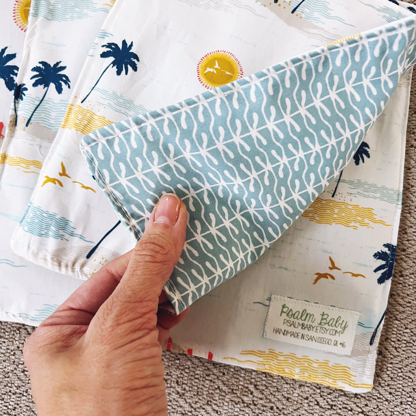 Hand holding a folded fabric item with palm tree and beach design, featuring a 'Psalm Baby' label.
