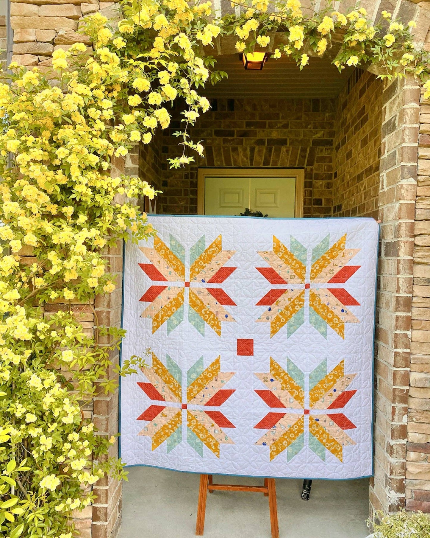 Quilt with star pattern displayed outdoors against a brick wall with yellow flowers.