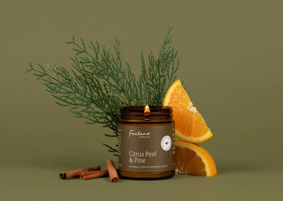 A lit brown glass candle jar labeled Fontana Citrus Peel & Pine, styled with orange slices, cinnamon sticks, and green pine branches against a muted olive-green background.