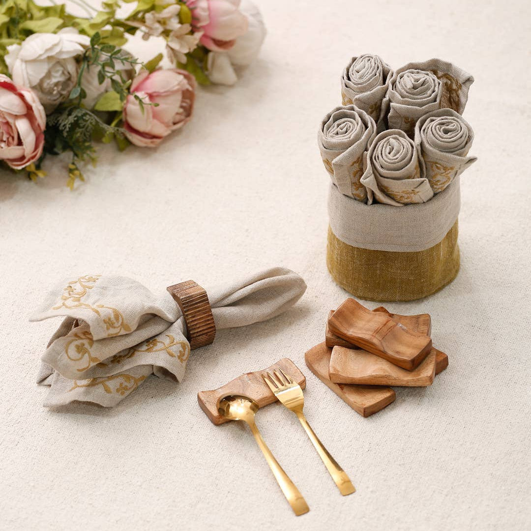 A styled flat-lay of neutral-toned tableware on a light beige textured surface, including rolled napkins in a fabric holder, a folded napkin with a brown ring, gold-toned forks on wooden coasters, and small wooden plates. Pink and white flowers appear in the upper left corner.