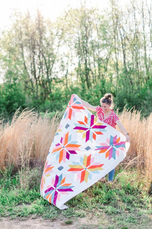 Person holding a colorful quilt in a natural setting with trees and tall grass.