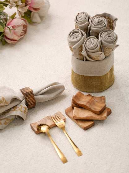 A styled flat-lay of a linen napkin set with gold cutlery and wooden cutters on a light beige textured surface, with a bundle of rolled napkins in a linen-wrapped holder and pink flowers in the background.